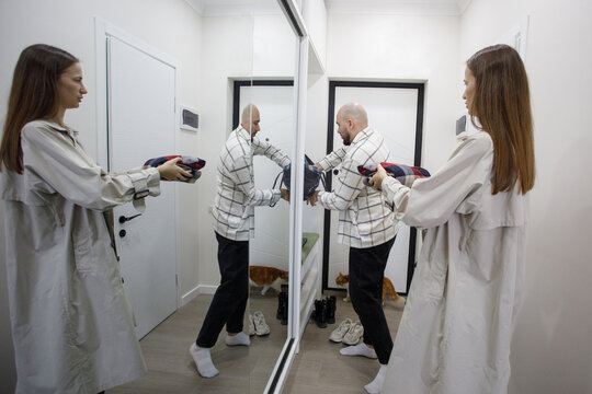 Person wearing a coat holds a hairdryer in front of a mirror while another person adjusts their outfit in a hallway