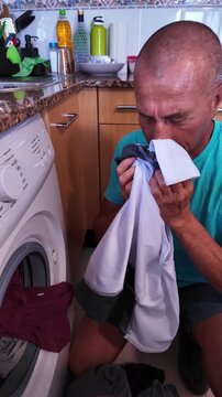 Middle-aged man smelling stinky, sweaty clothes with a disgusted expression on his face before putting them in the washing machine at home, performing a necessary but unpleasant household chore