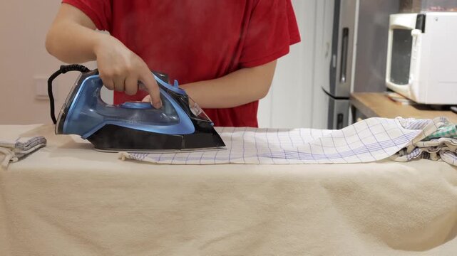 Hands iron a checked towel on a padded board in a home kitchen during a household laundry routine.