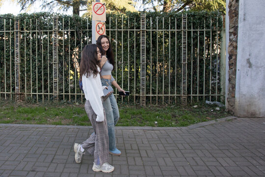 Mother with teenager  walk together on a sunny day along a path near tall green bushes and a fence in a city