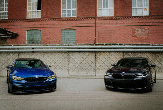 Front view of a blue BMW M4 Coupe and a dark purple BMW M5 Sedan parked together outdoors against a red brick building wall.
