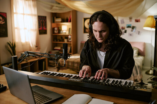 Young adult Caucasian man composing music on keyboard with headphones in home studio, producing audio content for independent artist branding, streaming, or music marketing