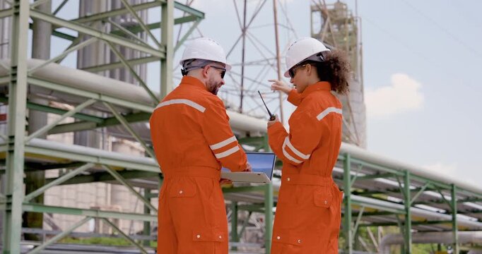 Asian female engineer uses walkietalkie beside multiracial male technician viewing laptop, updating refinery operation report, maintenance checklist, pipeline safety audit during site patrol