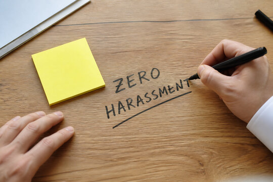 A hand writing ZERO HARASSMENT on a wooden desk next to a yellow sticky note.