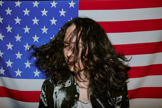 Young adult Caucasian man headbanging in front of United States flag, performing rock musician identity and youth culture concept. Useful for music promotion, patriotism campaigns