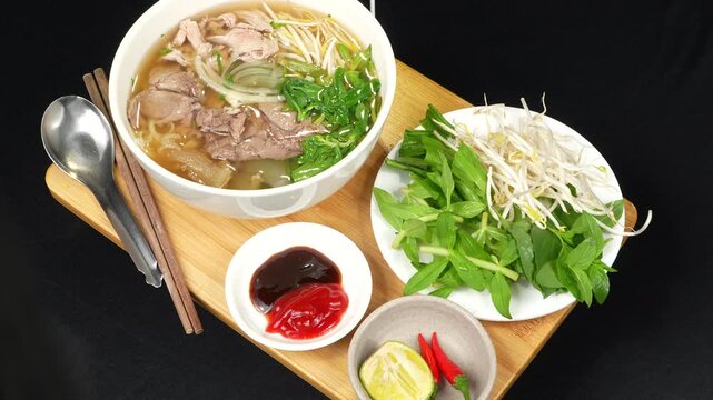 A complete Vietnamese Pho combo set on a wooden tray rotating, showing fresh herbs and sauces.