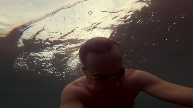 Swimmer with goggles looking up at the vibrant pink and orange sunset reflection from below the water surface