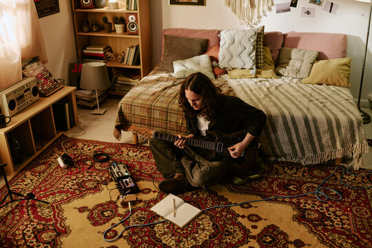 Young adult Caucasian man playing electric guitar on floor at home, practicing songwriting and music production. Useful for creative lifestyle, home studio, indie artist marketing