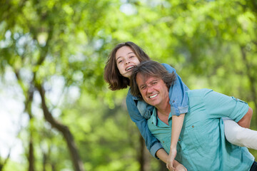 Fototapeta premium Cheerful father giving a piggyback ride to his joyful daughter in a sunny park, candid portrait of family connection and fun, lifestyle concept.