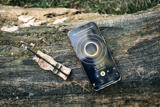 Smartphone displaying paused meditation app timer sits next to burning incense stick on weathered log. Pausing meditation session, taking a break app, mindful pause moment, Digital wellbeing feature.