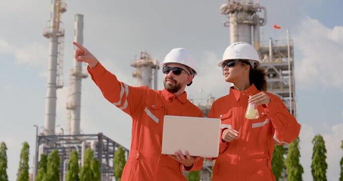 Multiracial male refinery technician points across processing tower while Asian female engineer carries sample flask, coordinating pipeline safety inspection route, maintenance compliance audit