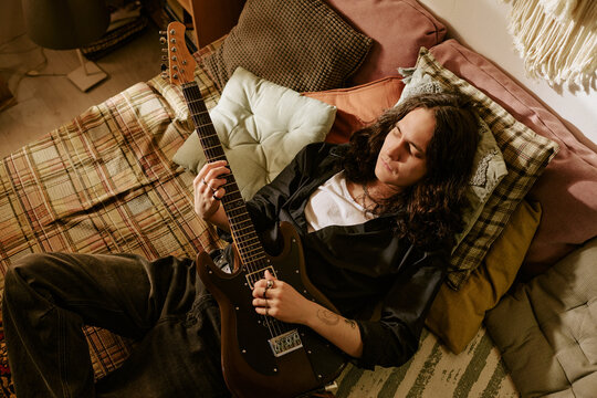 Young adult musician resting at home while holding electric guitar, expressing creative lifestyle and songwriting process. Useful for music branding, artist promotion, home studio content