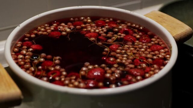 Wild strawberries and blackcurrants boiling in white ceramic pot on stove, homemade compote or jam cooking concept