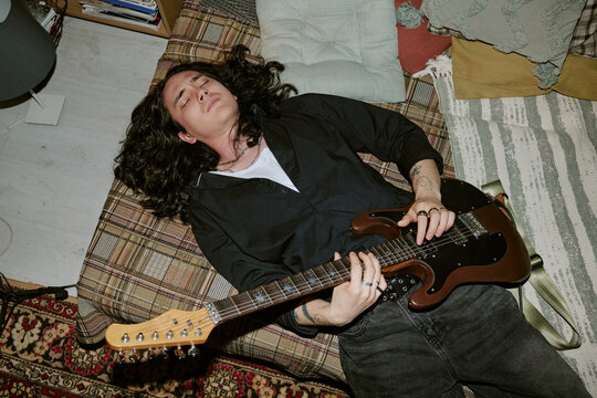 Young adult man playing electric guitar while lying on bed at home, expressing independent music lifestyle and creative passion. Useful for music promotion, artist branding, overhead shot