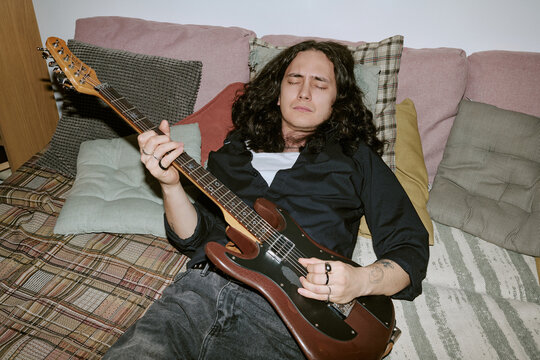 Young adult man playing electric guitar while reclining at home, expressing creative practice and independent musician lifestyle. Ideal for music lessons, artist branding, home studio marketing