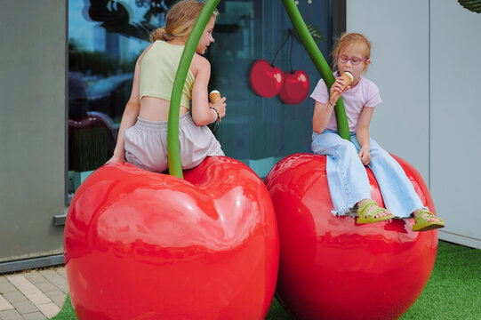 Girls enjoying ice cream on whimsical giant cherry playground sculptures on a sunny summer day