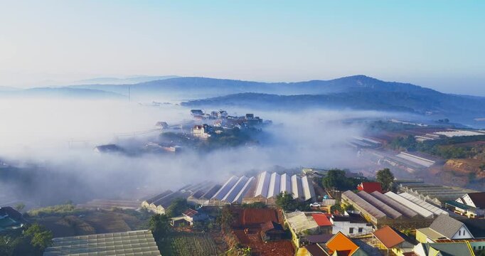 Aerial view of a small hillside village near Dalat, Vietnam wrapped in soft morning fog. Colorful houses emerge through layers of mist, while gentle sunlight illuminates rooftops and greenery.
