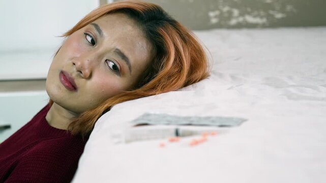 Close-up of a sad and lonely Asian woman leaning her head against a bed, looking off in despair with a medical syringe and scattered pills blurred in the foreground.