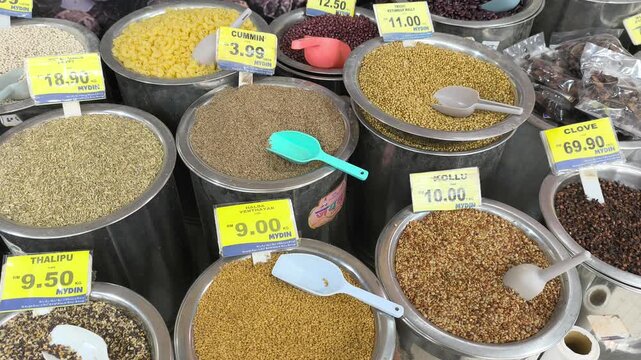 A variety of colorful spices are neatly displayed for sale in clean stainless steel jars. Each container features a clear price tag. The organized market setup looks hygienic and appealing.