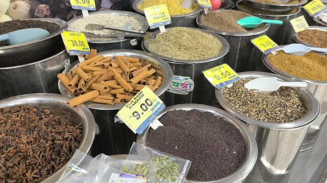 A variety of colorful spices are neatly displayed for sale in clean stainless steel jars. Each container features a clear price tag. The organized market setup looks hygienic and appealing.