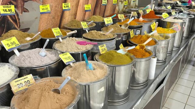 A variety of colorful spices are neatly displayed for sale in clean stainless steel jars. Each container features a clear price tag. The organized market setup looks hygienic and appealing.