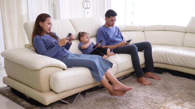 Young Asian family with a toddler sitting on a living room sofa, completely absorbed in their own individual smartphones and ignoring each other.