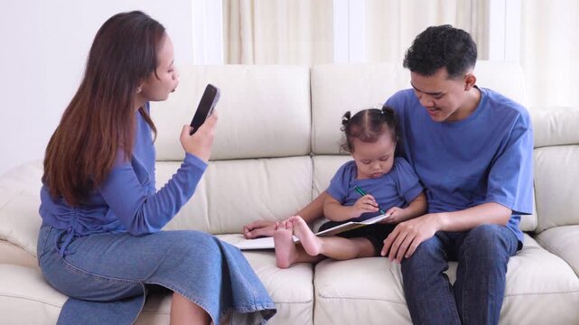 Asian mother using a smartphone to take a photo or video of her toddler daughter drawing in a notebook while sitting with her father on a sofa.