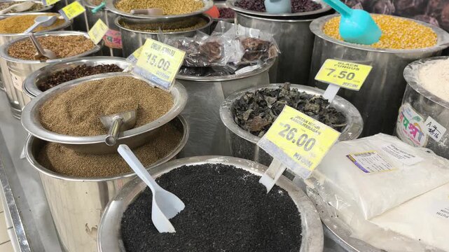 A variety of colorful spices are neatly displayed for sale in clean stainless steel jars. Each container features a clear price tag. The organized market setup looks hygienic and appealing.