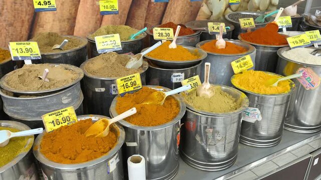 A variety of colorful spices are neatly displayed for sale in clean stainless steel jars. Each container features a clear price tag. The organized market setup looks hygienic and appealing.