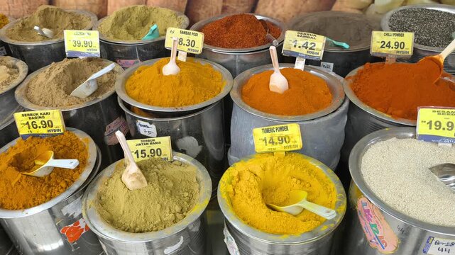 A variety of colorful spices are neatly displayed for sale in clean stainless steel jars. Each container features a clear price tag. The organized market setup looks hygienic and appealing.