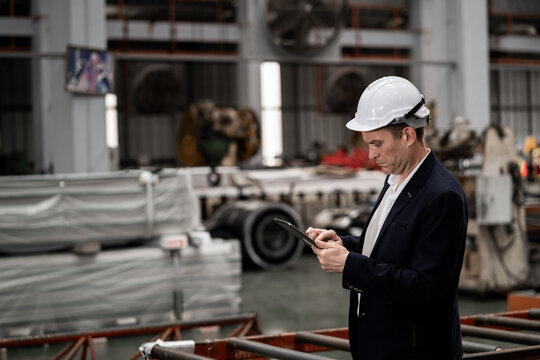 Male Manager in Suit and White Hard Hat Using Tablet at Steel Manufacturing Factory. Business Professional Conducting Site Inspection in Heavy Industry.