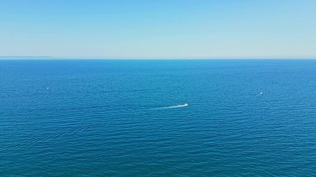 A boat moves across a wide blue ocean, creating gentle waves on the surface. The sky is clear and sunny, with no clouds present.