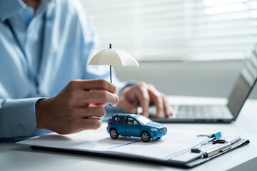 Fototapeta premium Insurance agent holding umbrella over a blue toy car while working on a laptop computer, focusing on vehicle protection, digital finance, and online safety