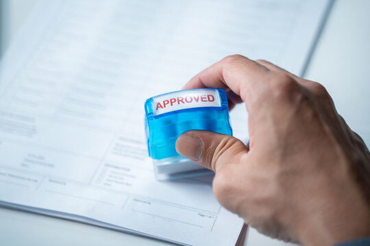 Businessman hand pressing a stamp with the word approved on a white paper document, representing agreement finalization and official loan acceptance