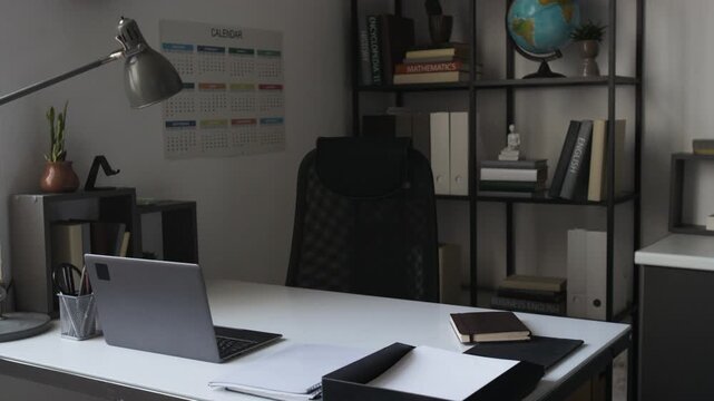 Empty teachers desk holding laptop, notebook, and lamp with bookshelf in background. Footage for back-to-school marketing campaigns or educational promo videos