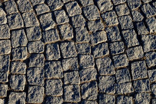Granite cobblestone pavement texture with curved pattern. Rough stone paving block surface under bright low sun with contrast shadow and hard side lighting, close up.