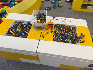 Fototapeta premium Lego bricks sorted in plastic bins on a table in a playroom, with colorful toy pieces and a children’s play area in the background