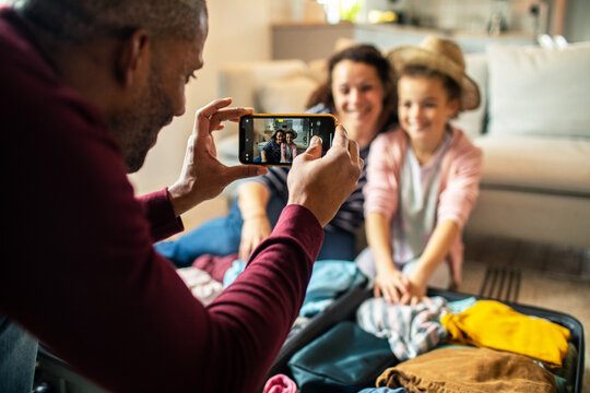 Father photographing mother and daughter packing suitcase at home for vacation