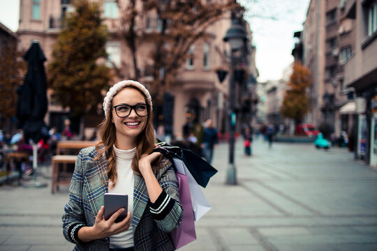 Smiling young woman with shopping bags using smartphone on city street