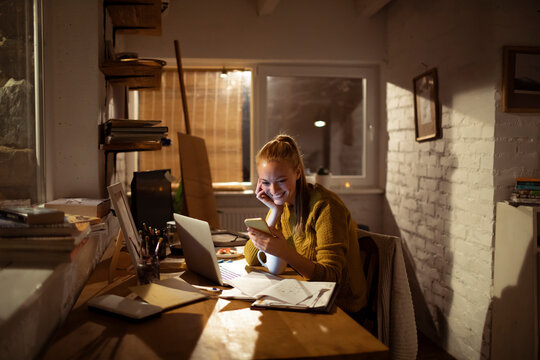 Young woman working on laptop and phone in cozy home office at night