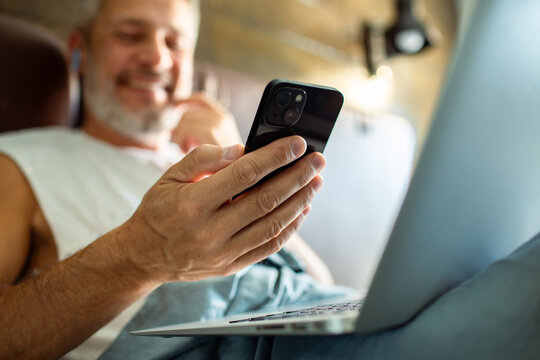 Smiling mature man using smartphone and laptop at home