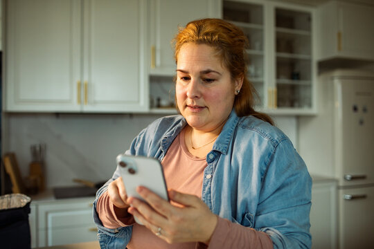 Thoughtful woman using smartphone in modern home kitchen