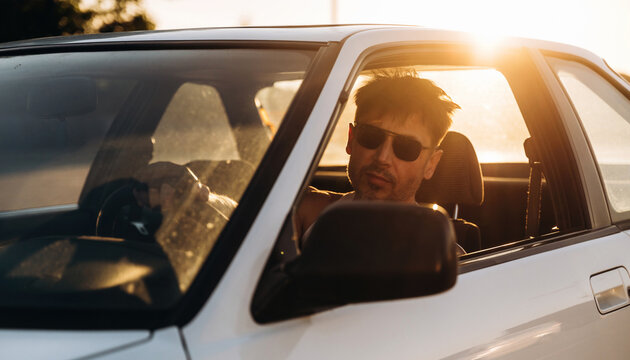 Man wearing sunglasses sits in a white car with sunlight streaming through the windshield, showcasing a relaxed atmosphere during a sunny day