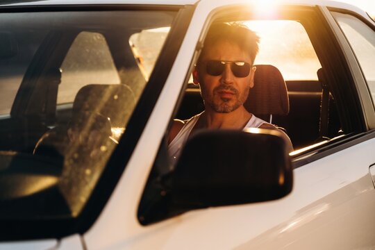 Male driver wearing sunglasses sits in a white car with sunlight reflecting through the window, showcasing a relaxed demeanor and urban environment in the background