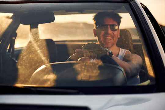 Male driver wearing sunglasses smiles while holding steering wheel of a car, sunlight streaming through windshield, with blurred landscape visible in the background
