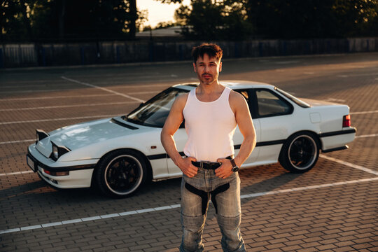 Male car enthusiast in white tank top and jeans poses confidently beside modified white sports car in empty parking lot during sunset