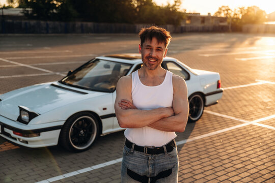 Caucasian male car enthusiast stands confidently with arms crossed in front of a white sports car parked on a sunlit asphalt lot during golden hour