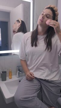 Young woman in cozy pajamas performs a home beauty routine in the bathroom using a microcurrent facial device for a relaxing face massage and daily skincare ritual.