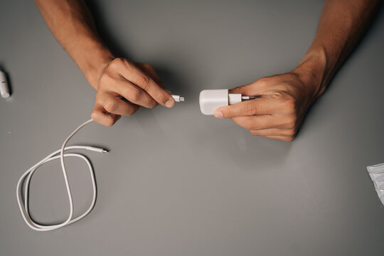 Close-up top view of man connecting USB cable to electronic cigarette charger on gray desk, showcasing charging process of vaping device. Concept of modern alternative to traditional smoking.