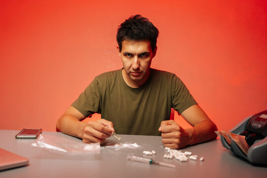 Front view of drug dealer preparing to snort cocaine with rolled banknote sitting at table with pills and syringe on red background, looking at camera, representing drug addiction and substance abuse.